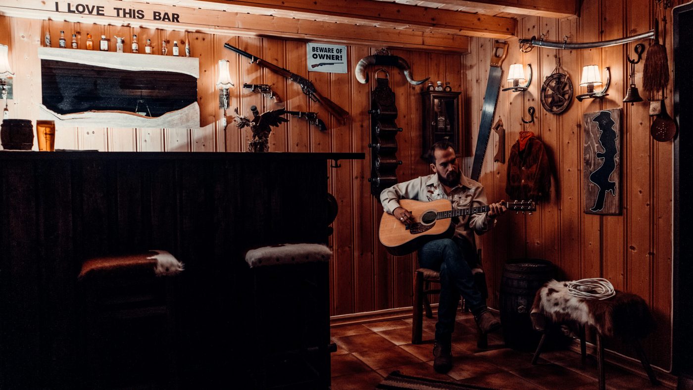 Johan med gitar på en krakk i en bar med tømmervegger.